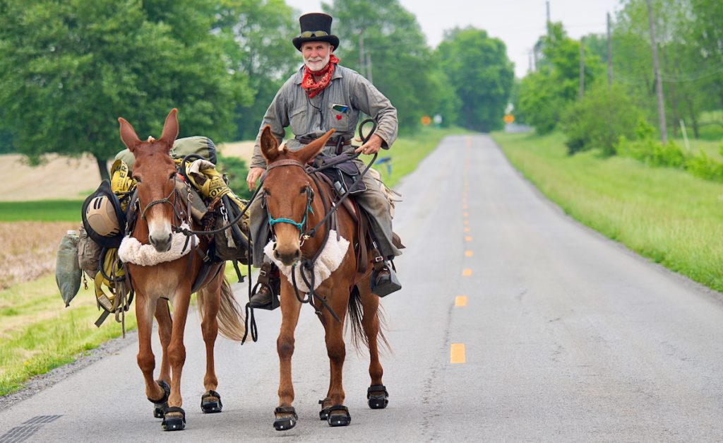 Long Rider Bernie Harberts riding mule Cracker and leading pack mule Brick down a country road, Two Mules to Triumph